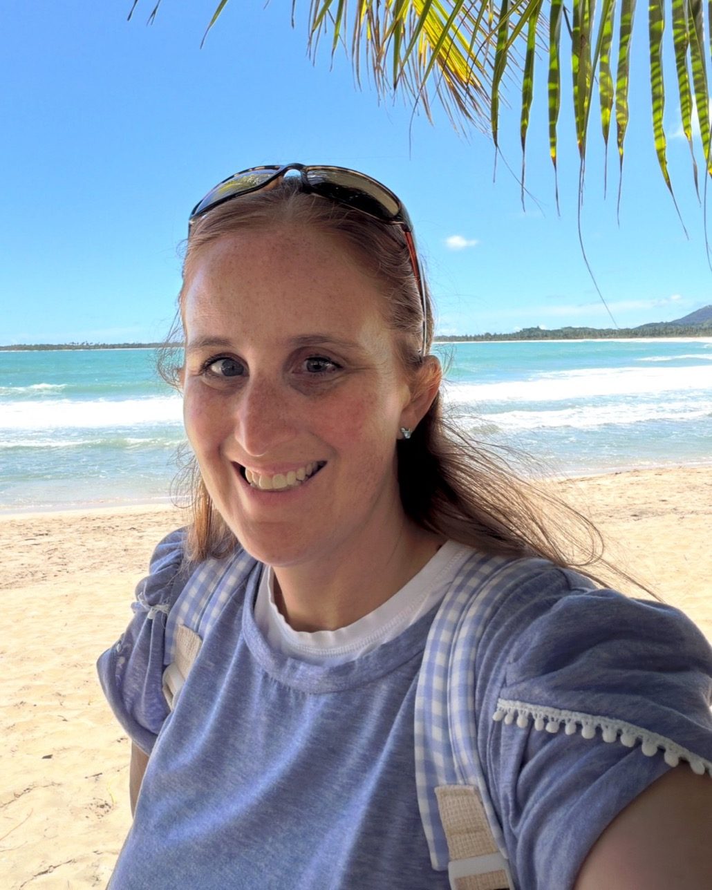 Melanie Francis smiling on a tropical beach beneath a palm frond